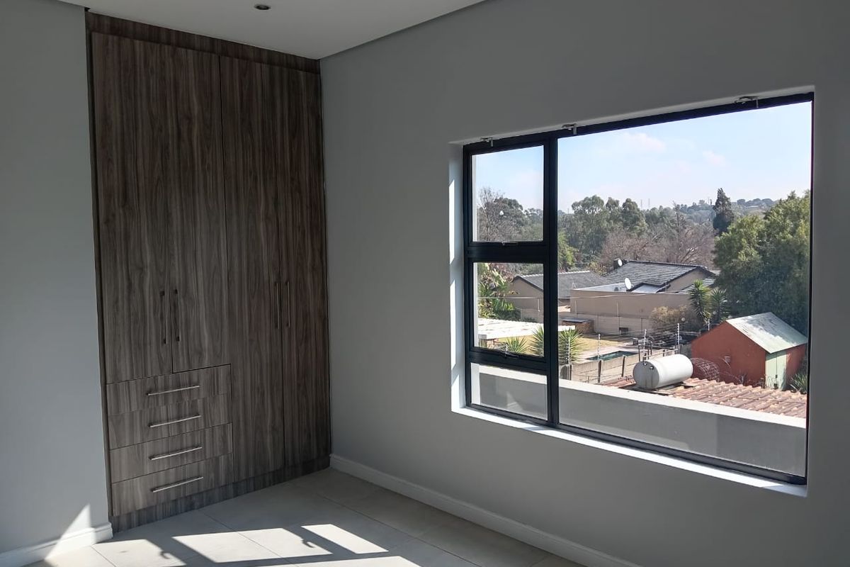 A bright, empty bedroom featuring modern wood-finish built-in cupboards, tiled flooring, and a large window offering an elevated neighbourhood view.