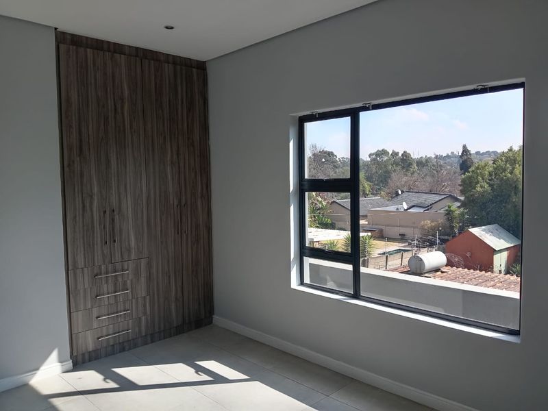 A bright, empty bedroom featuring modern wood-finish built-in cupboards, tiled flooring, and a large window offering an elevated neighbourhood view.