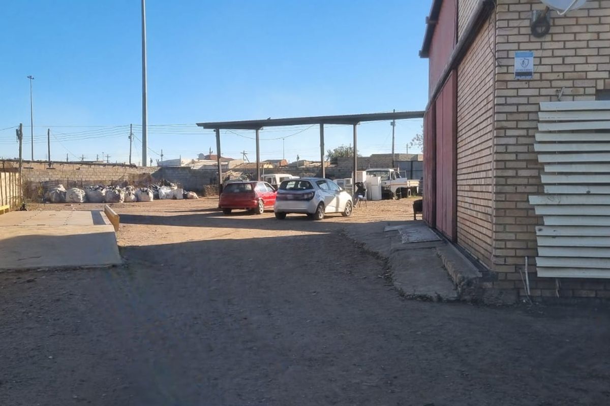 Rear exterior view of a commercial property featuring a brick facade, concrete loading ramp, and a metal carport structure over an unpaved yard.