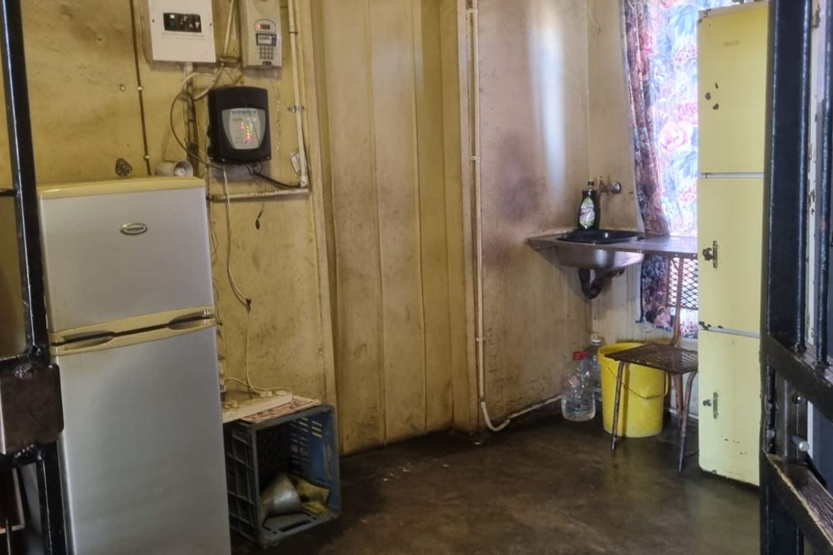 A small, utilitarian kitchen area featuring a wall-mounted sink, security gate access, and significant wear on the wall surfaces requiring refurbishment.