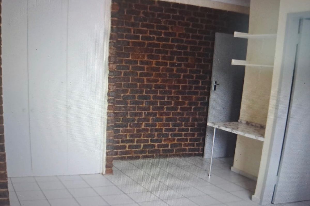 An empty living area featuring an exposed brick feature wall, tiled flooring, and built-in shelving with a small counter space.