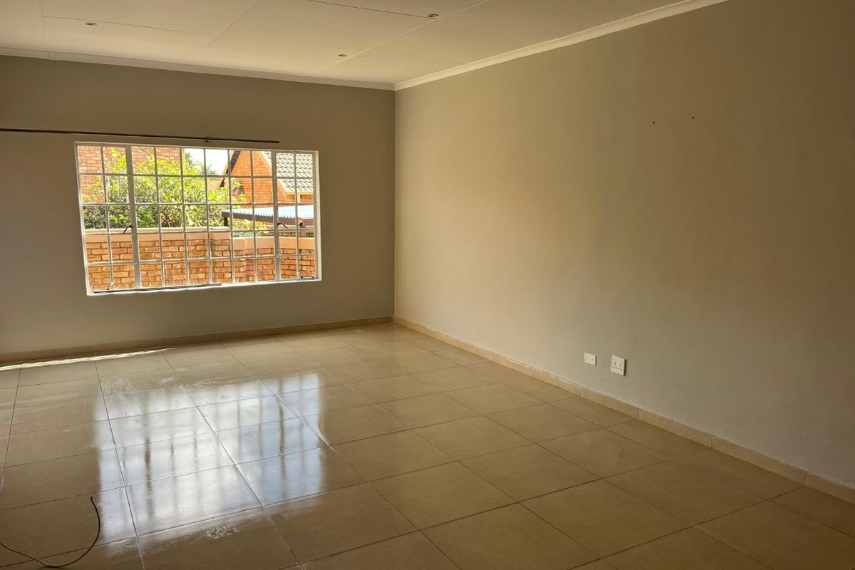 A spacious, empty living area featuring neutral wall tones, tiled flooring, and a large grid-pattern window providing natural light.