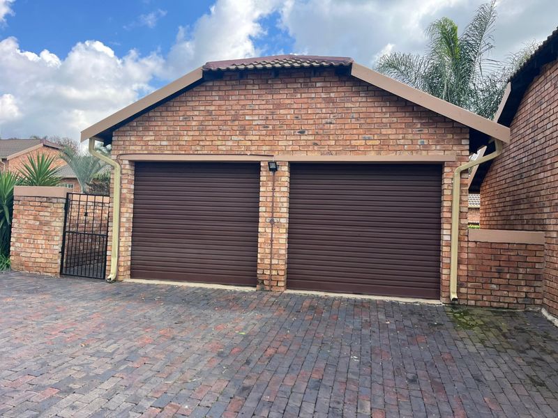 A double garage featuring face brick construction, brown roller doors, and a paved driveway, set within a secure townhouse complex.