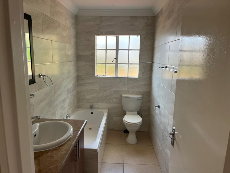A clean, neutral-toned bathroom featuring a built-in vanity, bathtub, and toilet with modern wall and floor tiling.