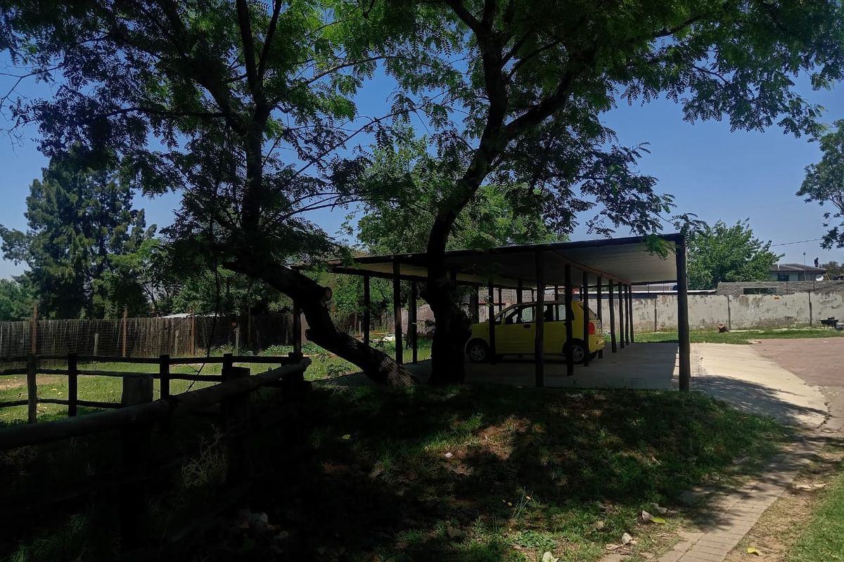 A functional outdoor carport structure providing covered parking, situated on a residential plot with established trees and perimeter fencing.