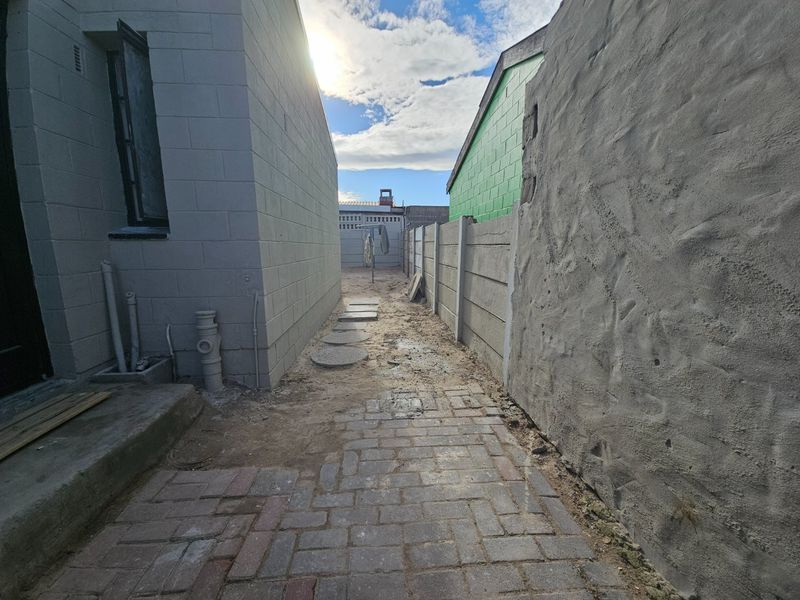 A narrow side passage featuring paved walkways, concrete block walls, and exposed external plumbing infrastructure.