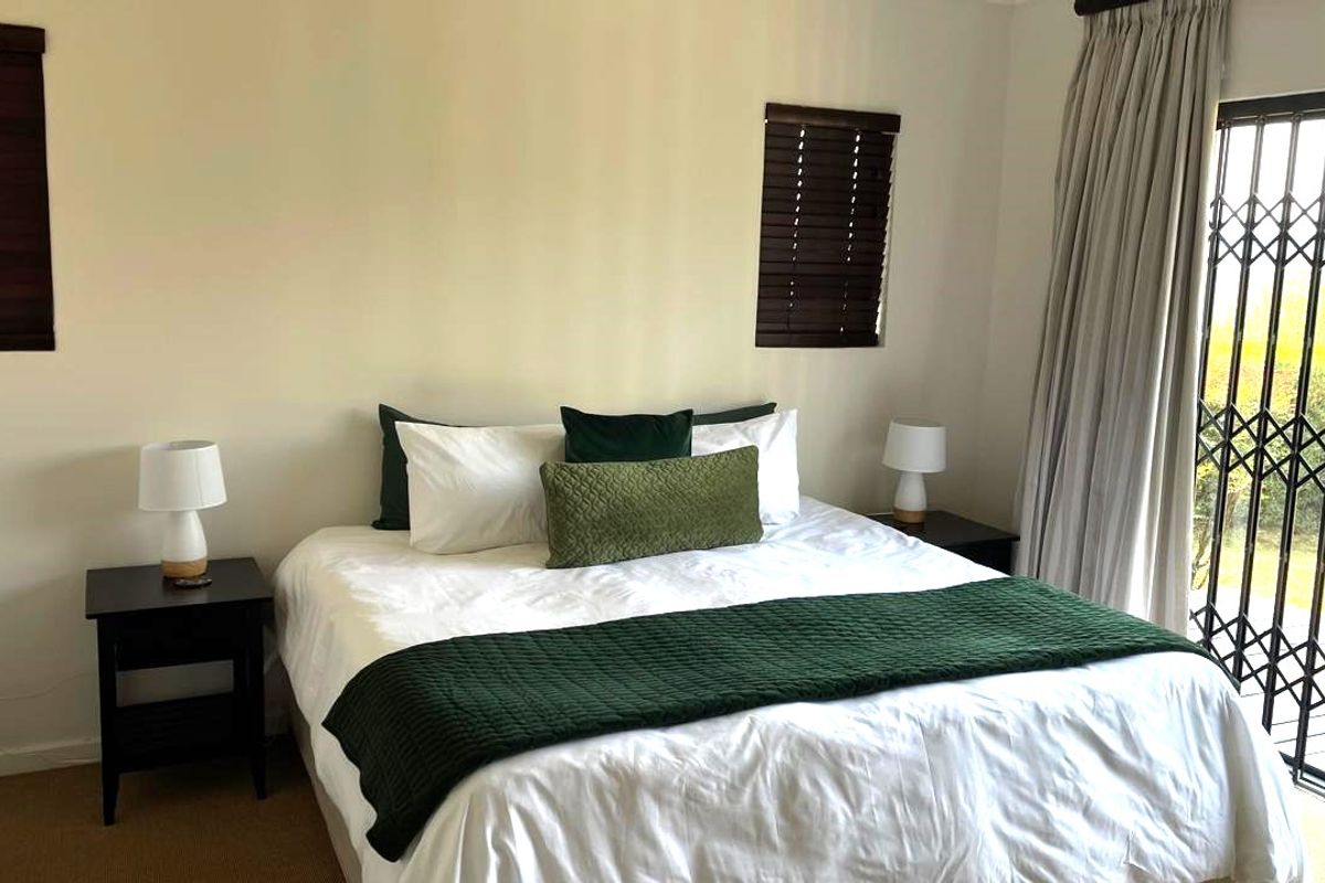 A well-lit bedroom featuring neutral walls, wooden blinds, and a sliding door with a security gate leading to the exterior.
