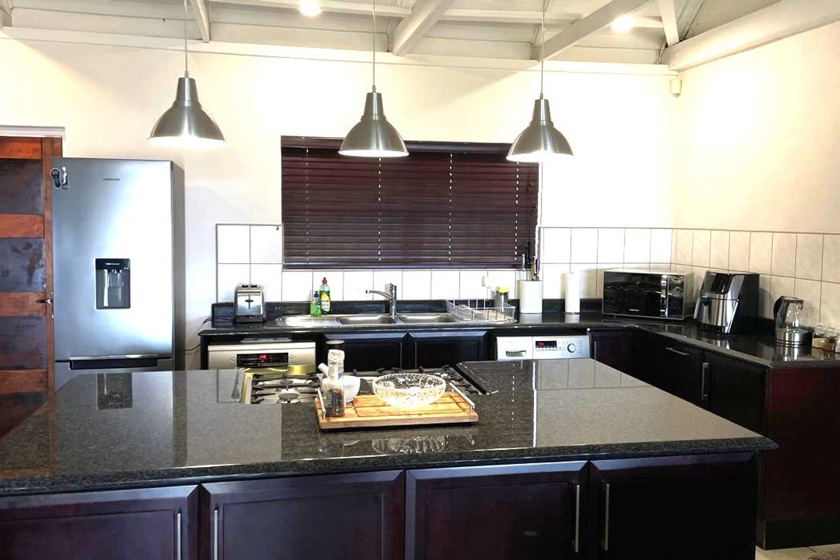 A functional kitchen featuring a large central island with a granite countertop, built-in gas hob, and modern pendant lighting under an exposed beam ceiling.