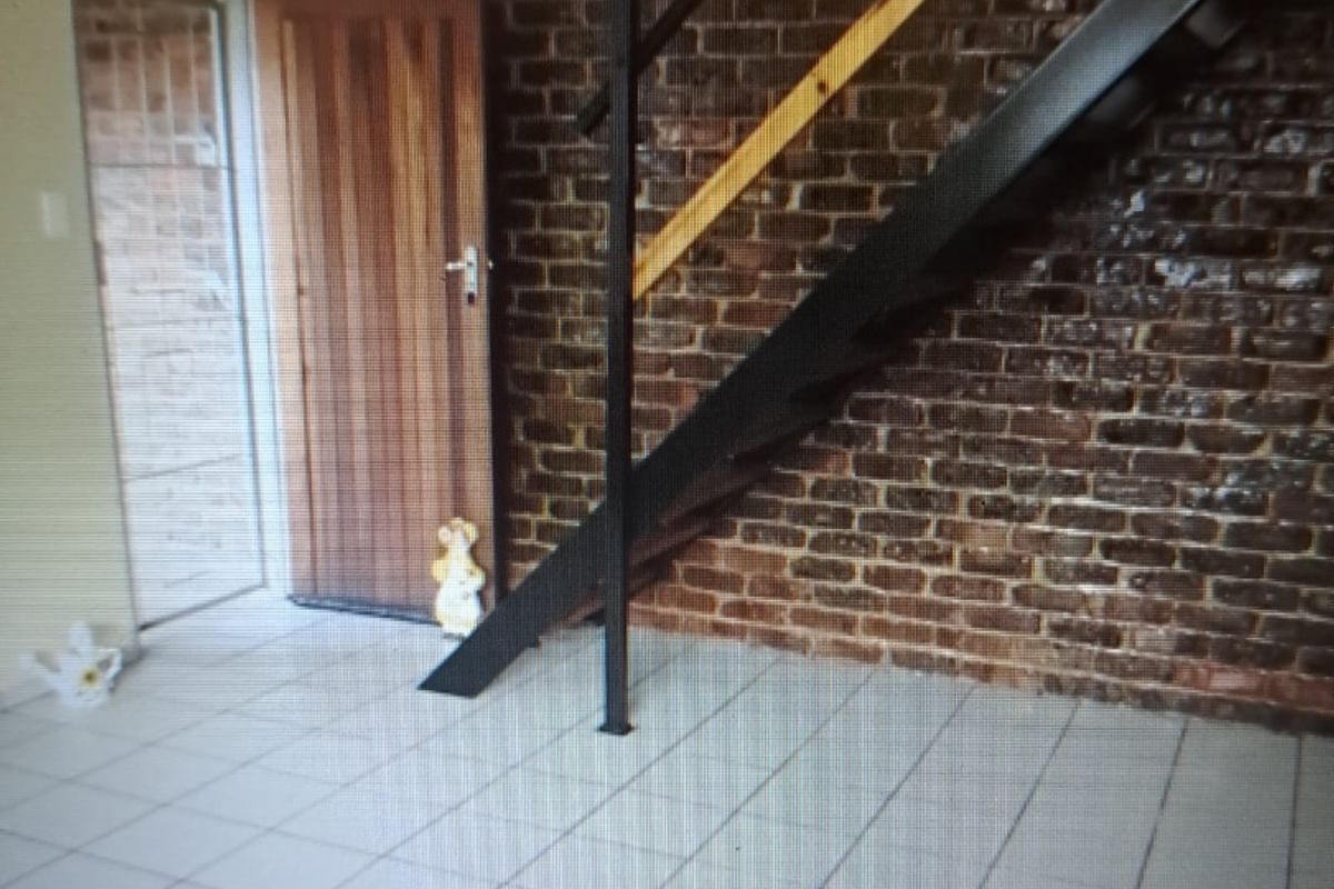 An open-plan living area featuring an exposed brick feature wall, ceramic floor tiles, and a modern metal staircase leading to the upper level.