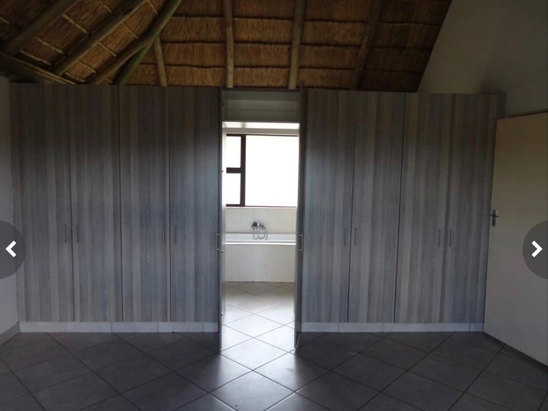 Spacious bedroom featuring extensive built-in cupboards, high thatched ceiling, and tiled flooring, with an adjoining bathroom visible through the central doorway.