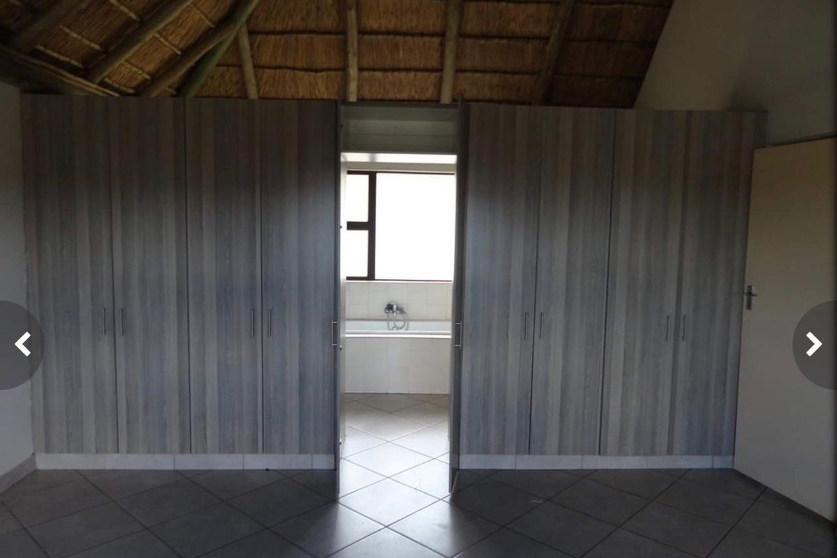 Spacious bedroom featuring built-in cupboards, tiled flooring, and a traditional thatched roof, with an adjoining bathroom visible through the doorway.