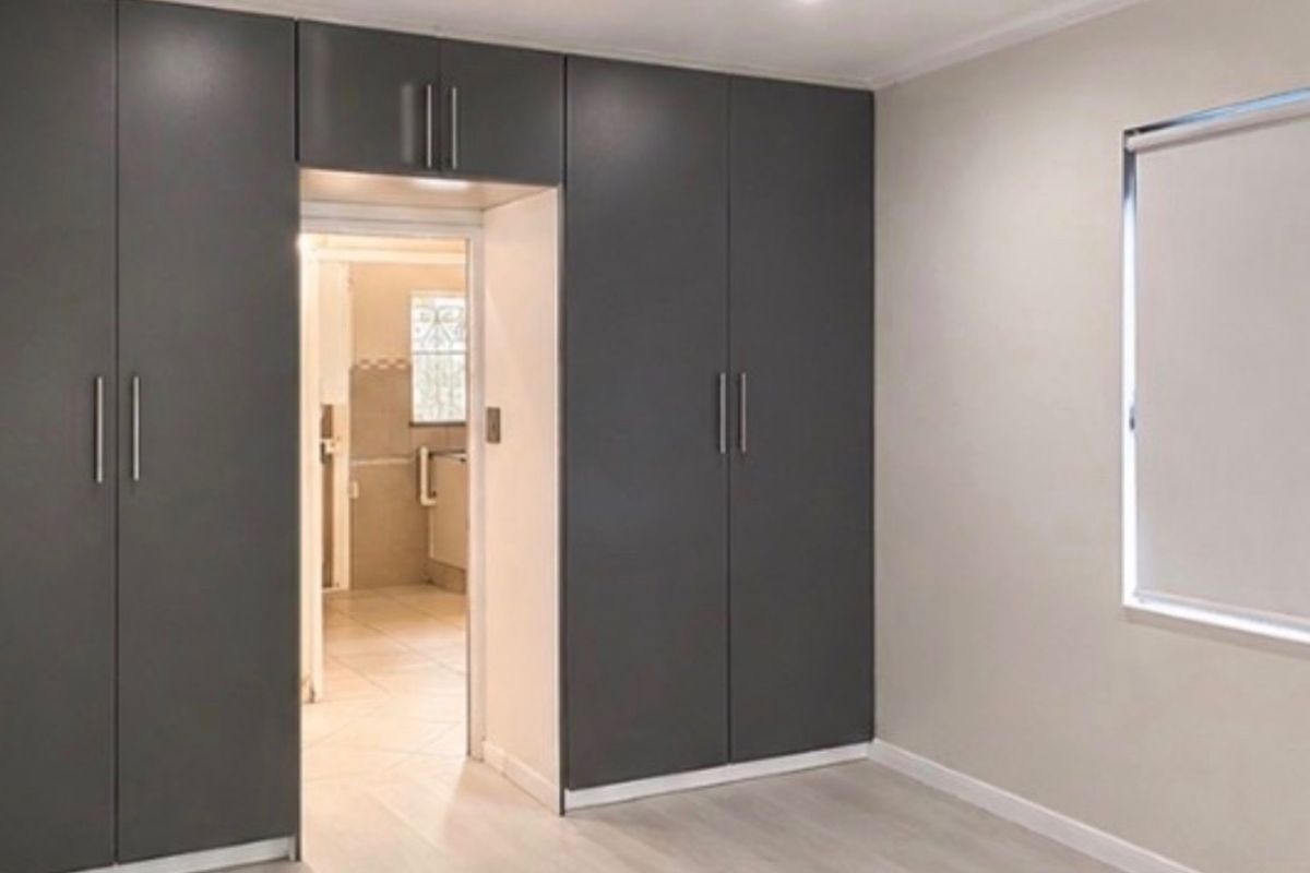 A clean, modern bedroom featuring dark grey built-in cupboards, light laminate flooring, and a roller blind, with an adjoining bathroom visible.