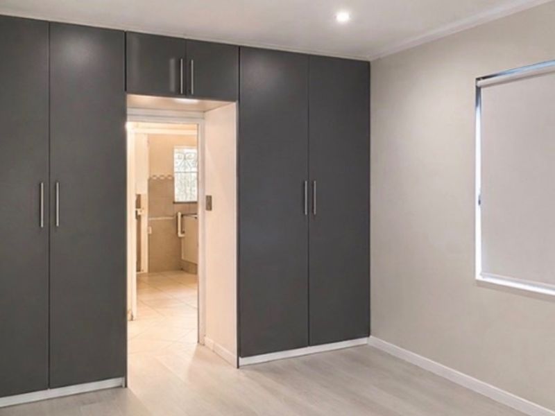 A clean, modern bedroom featuring dark grey built-in cupboards, light laminate flooring, and a roller blind, with an adjoining bathroom visible.