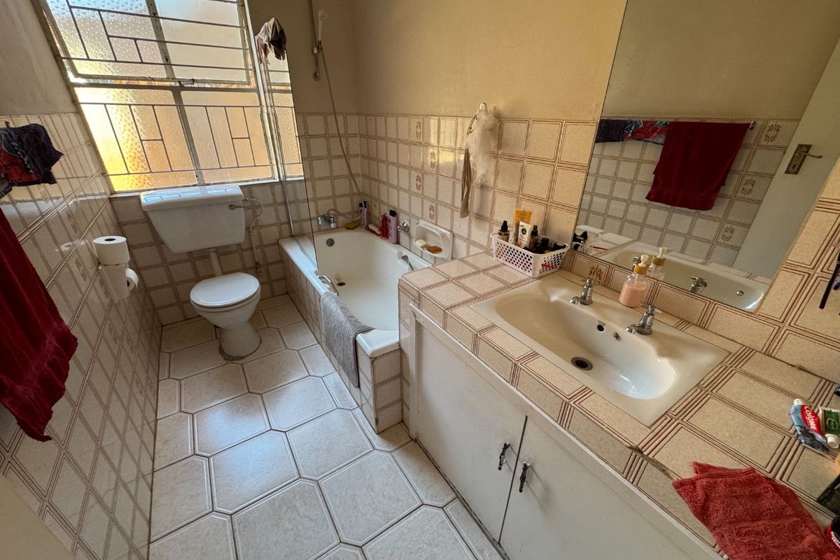 A dated bathroom featuring a built-in vanity, bathtub, and toilet with original tiled finishes and security bars on the window.
