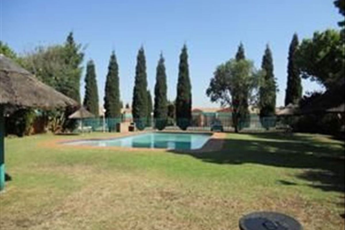 Communal outdoor area featuring a swimming pool, manicured lawn, and established trees within a fenced complex setting.