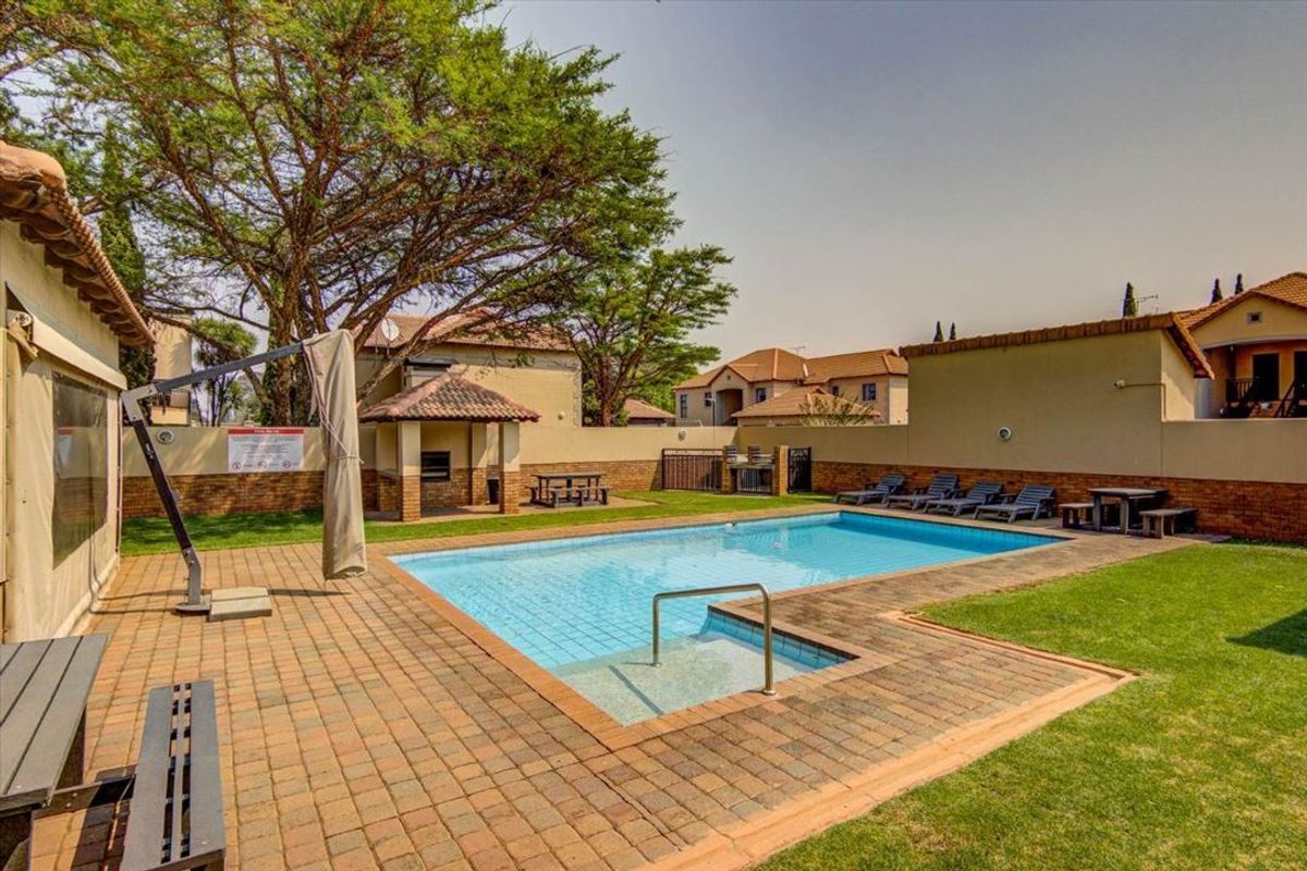 Communal pool area featuring a paved deck, built-in braai facility, and surrounding lawn within a secure townhouse complex.