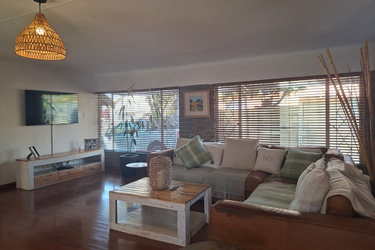 A comfortable living room featuring warm timber flooring, an exposed brick accent wall, and large windows fitted with venetian blinds.