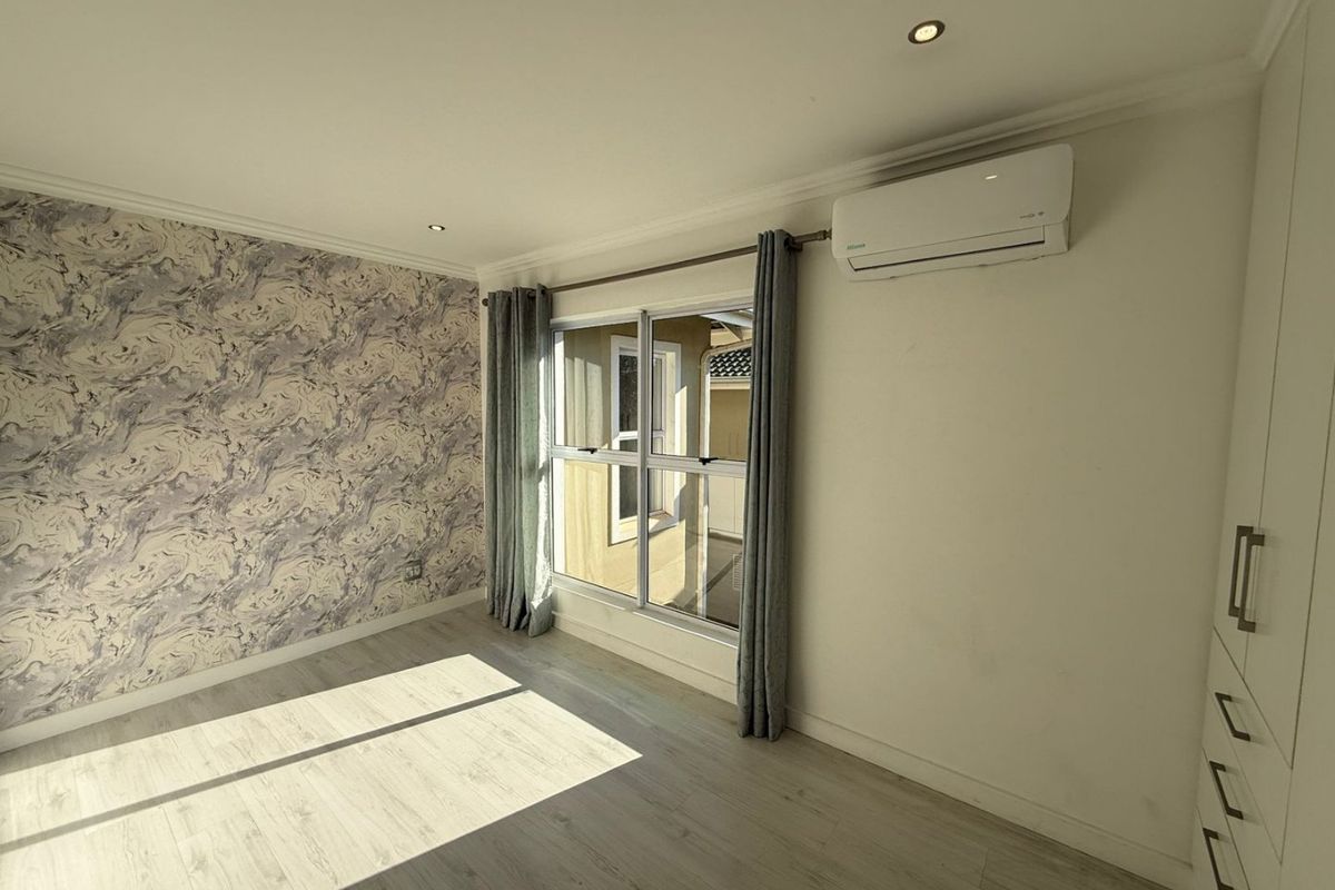 A bright, empty bedroom featuring modern laminate flooring, built-in cupboards, an air conditioning unit, and a feature wallpaper wall.