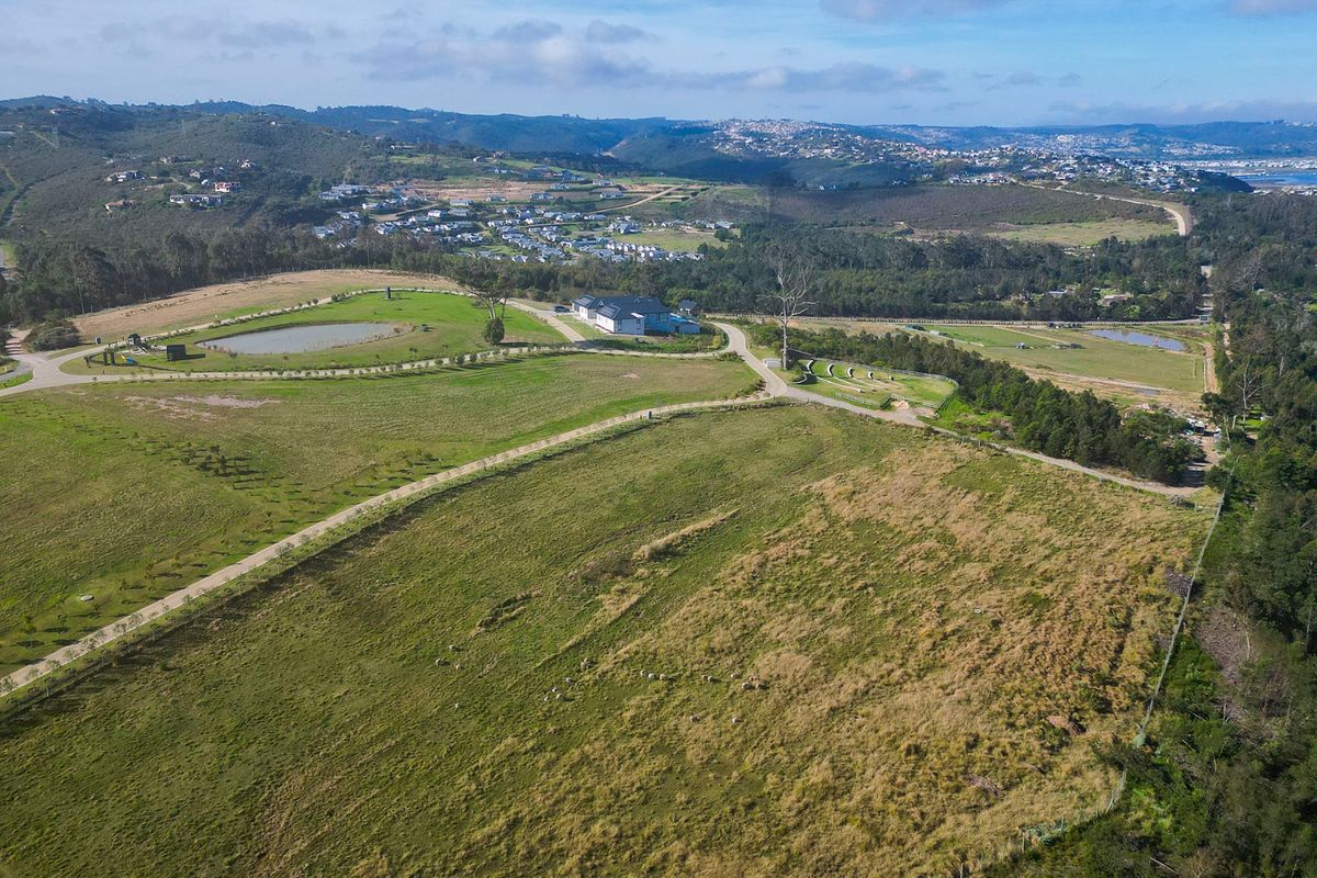 An aerial view of a large residential land plot featuring rolling green hills, paved access roads, and a small dam.