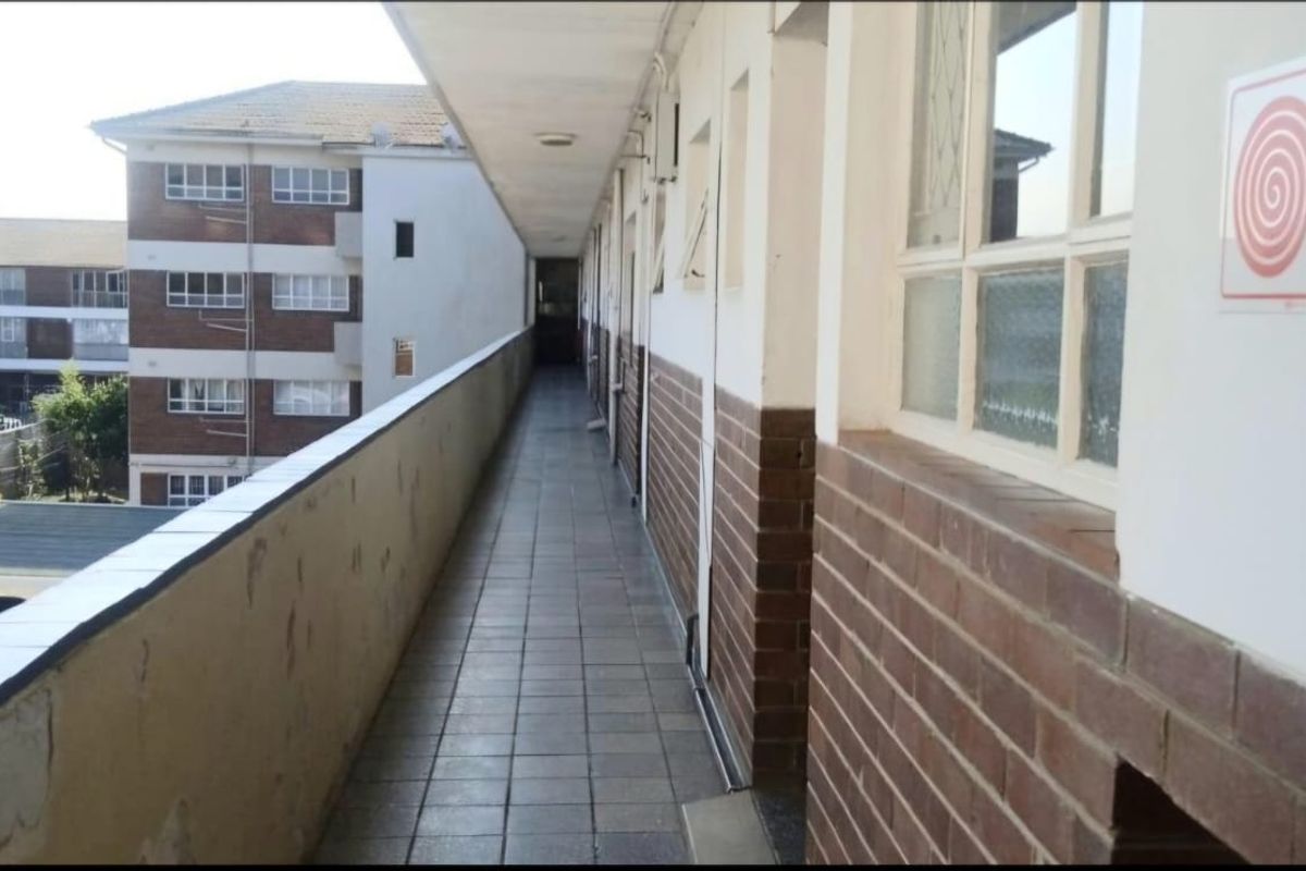 An exterior apartment building walkway featuring tiled flooring, brick wall finishes, and safety signage, providing access to residential units.