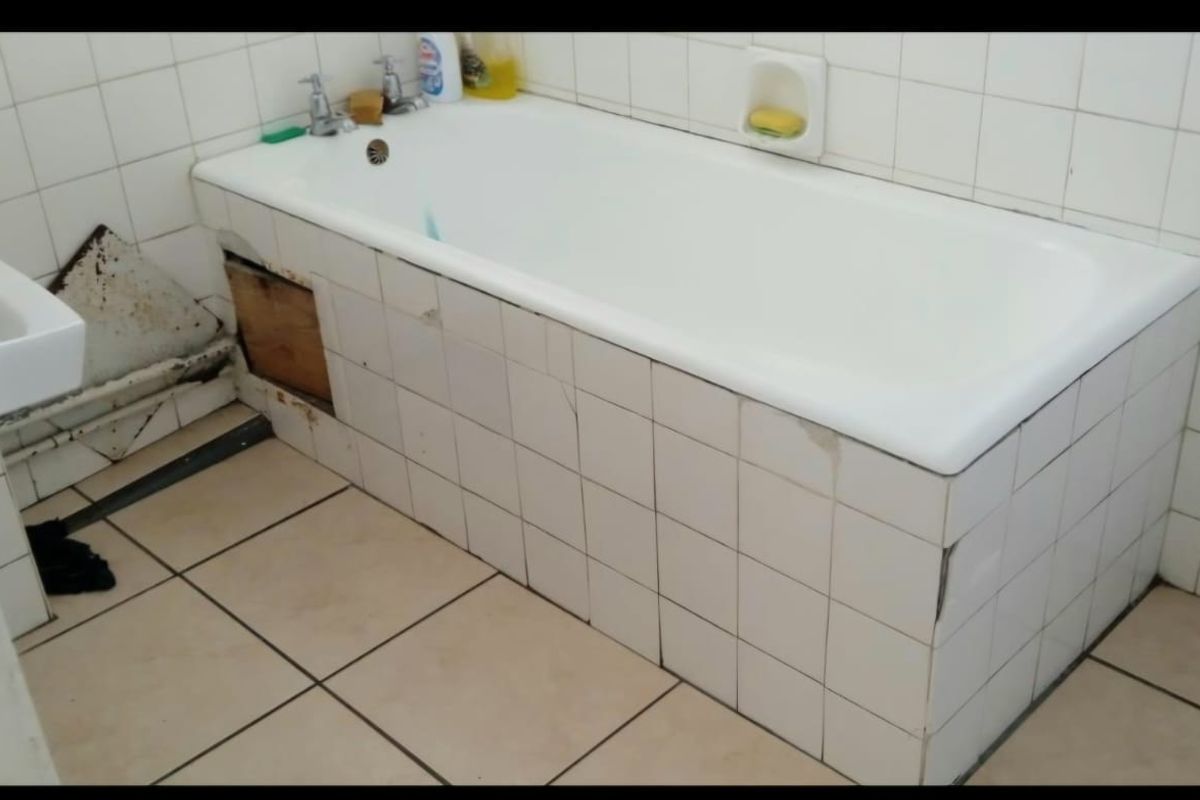 A dated bathroom featuring a tiled bathtub with visible structural damage and missing paneling, requiring significant renovation and repair to the tiling and fixtures.