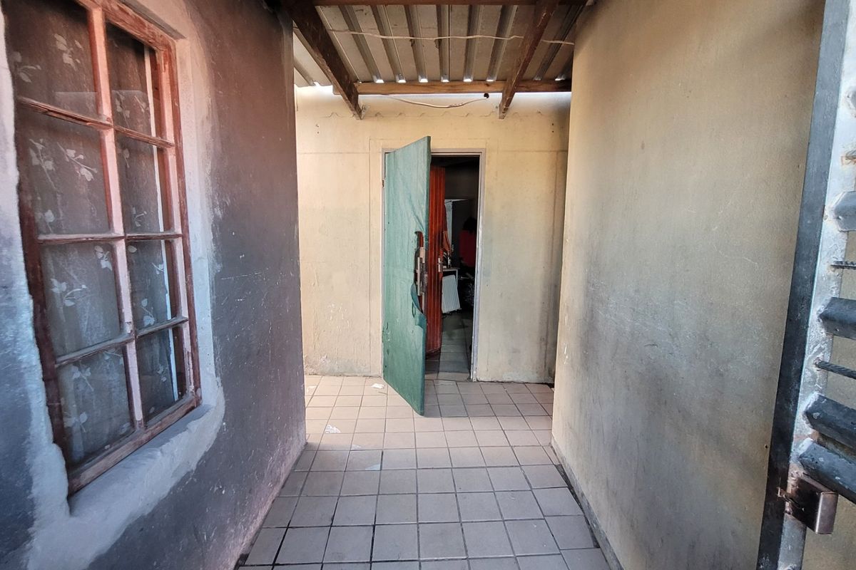A covered outdoor walkway or patio area featuring tiled flooring and corrugated roofing, showing signs of wear and requiring maintenance.