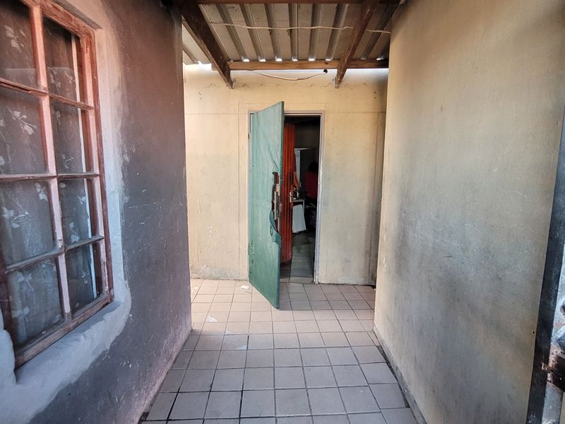 A covered outdoor walkway or patio area featuring tiled flooring and corrugated roofing, showing signs of wear and requiring maintenance.
