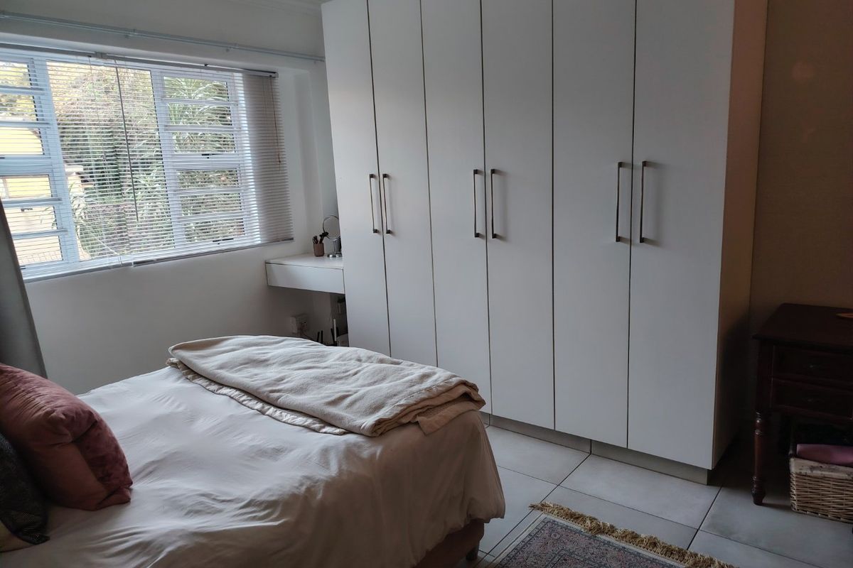 A well-maintained bedroom featuring extensive white built-in cupboards, neutral tiled flooring, and a window with fitted blinds providing natural light.