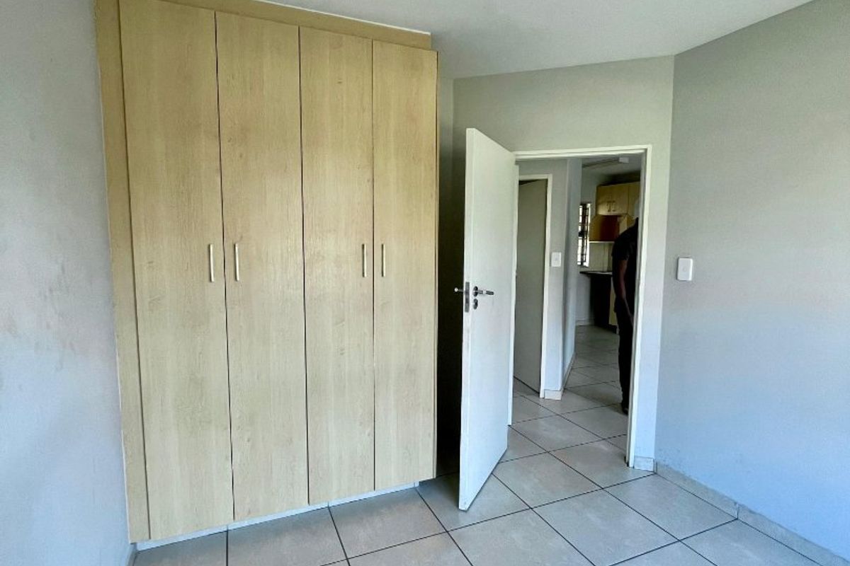 A vacant bedroom featuring neutral tiled flooring and built-in wooden cupboards, with a view through the doorway into the adjacent kitchen area.