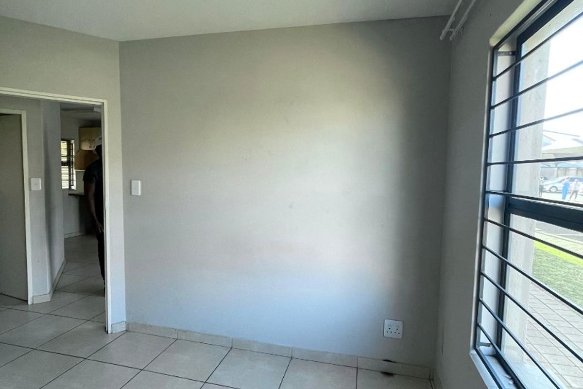 An empty living area featuring neutral tiled flooring, a window with security bars, and access to an adjacent room.