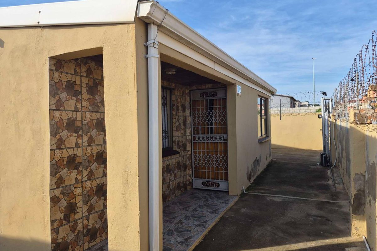 Rear exterior view of the property featuring a covered entrance, security gates, and concrete paving, enclosed by a perimeter wall with razor wire.