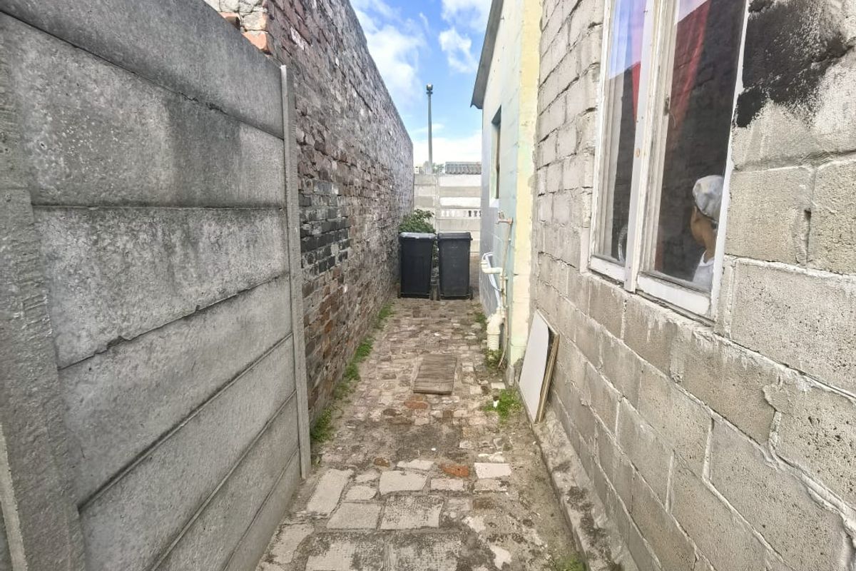 A narrow side passage featuring a concrete block boundary wall, exposed brickwork, and weathered paving stones leading to the rear of the property.
