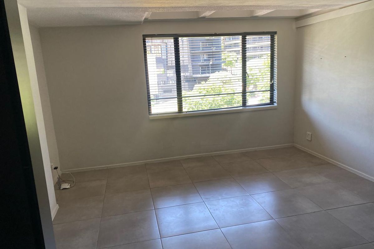 A bright, empty living room featuring large format floor tiles, neutral walls, and a large window with venetian blinds providing natural light.