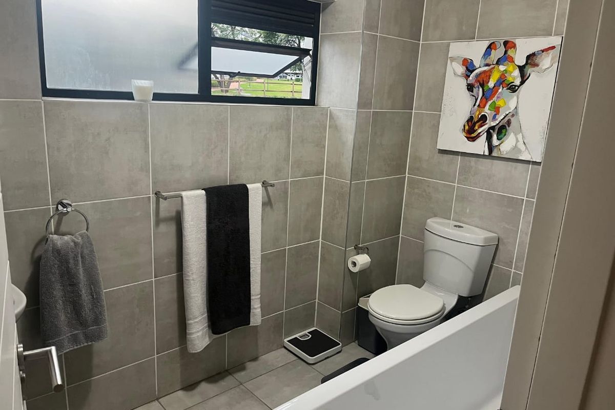 A modern bathroom featuring neutral grey wall and floor tiles, a white bathtub, and a toilet, complemented by a top-hung window for natural light.