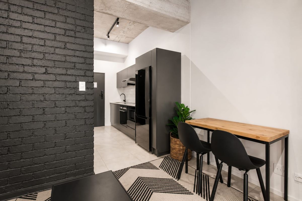 Modern open-plan apartment featuring a sleek kitchen with dark cabinetry, exposed concrete ceilings, and a striking black brick feature wall.