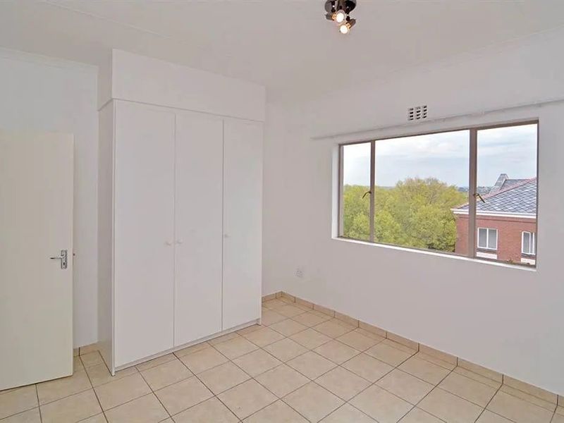 A bright, empty bedroom featuring neutral tiled flooring, built-in cupboards, and a large window providing natural light and an elevated view.