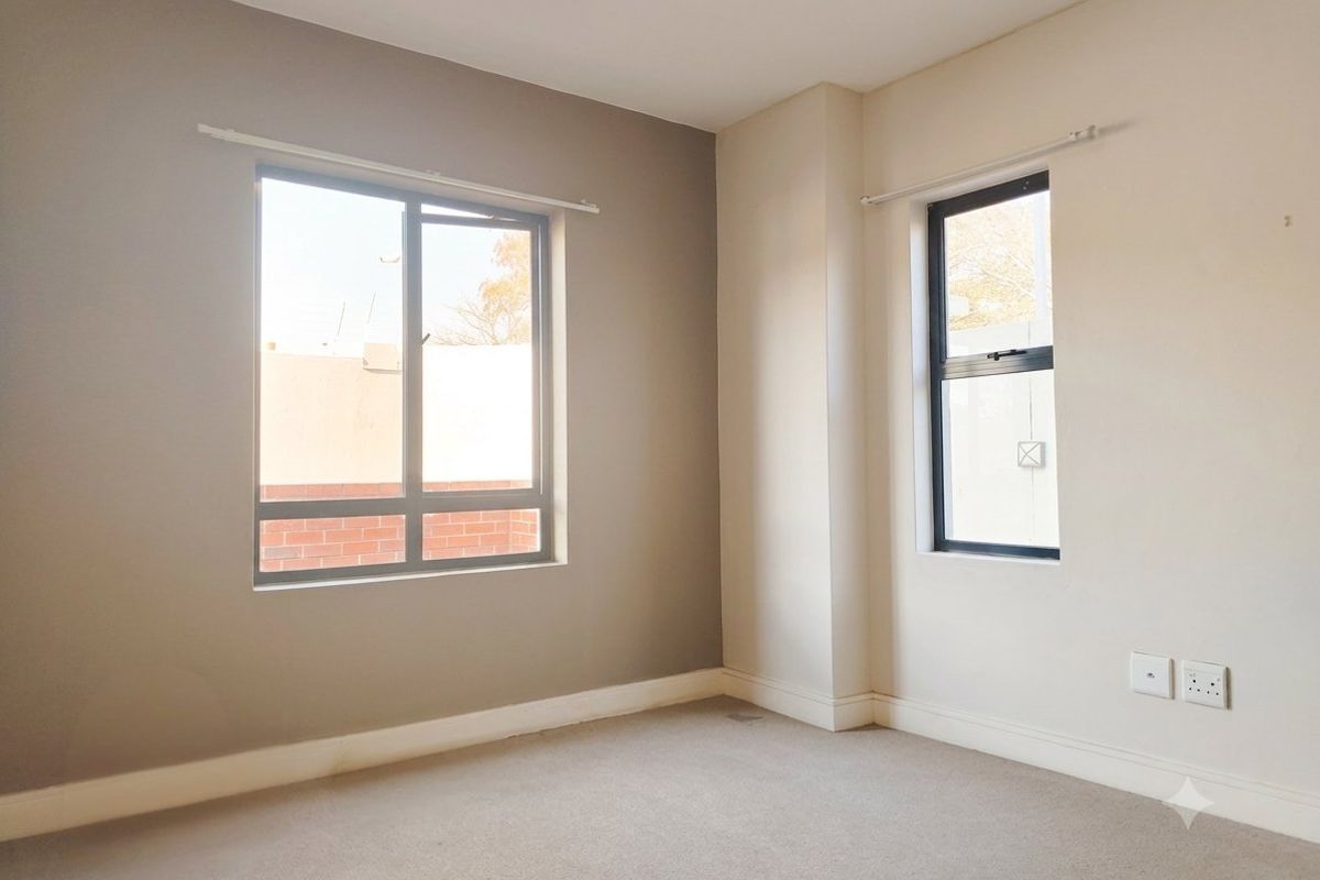 A bright, empty bedroom featuring neutral wall tones, carpeted flooring, and aluminium-framed windows providing natural light.