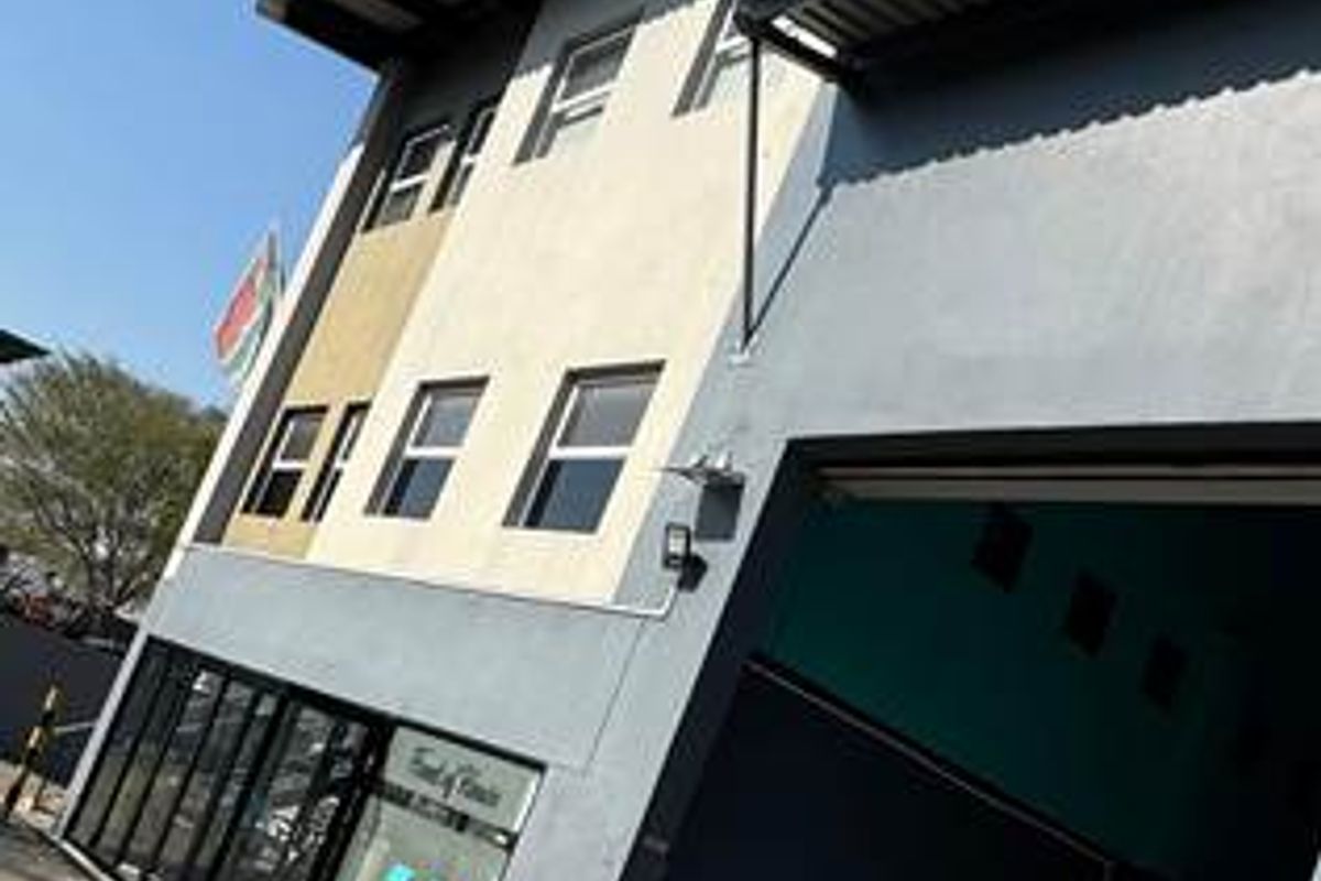 A modern commercial building featuring a large roller shutter door, glass storefront entrance, and a metal roof, situated on a paved driveway.