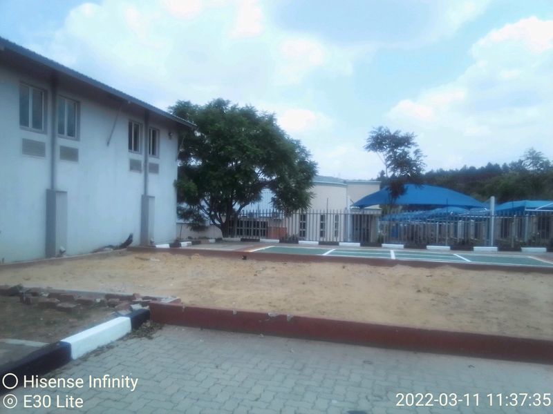 Rear exterior view of the commercial property featuring a paved driveway, perimeter fencing, and a shaded outdoor area with unfinished ground surfacing.