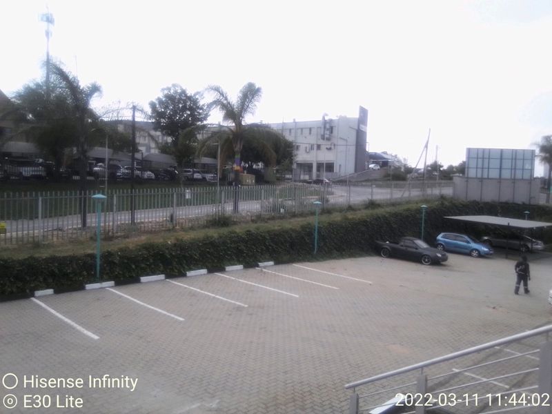 An elevated view of a commercial paved parking area featuring a carport, security fencing, and landscaped perimeter hedging.