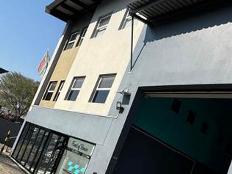 A modern commercial building featuring a large roller shutter door, glass storefront entrance, and a metal roof, situated on a paved driveway.