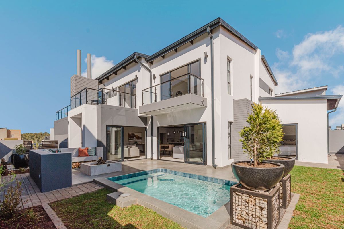 Modern double-story rear elevation featuring a swimming pool, paved patio area, built-in braai, and upper-level balconies with glass balustrades.