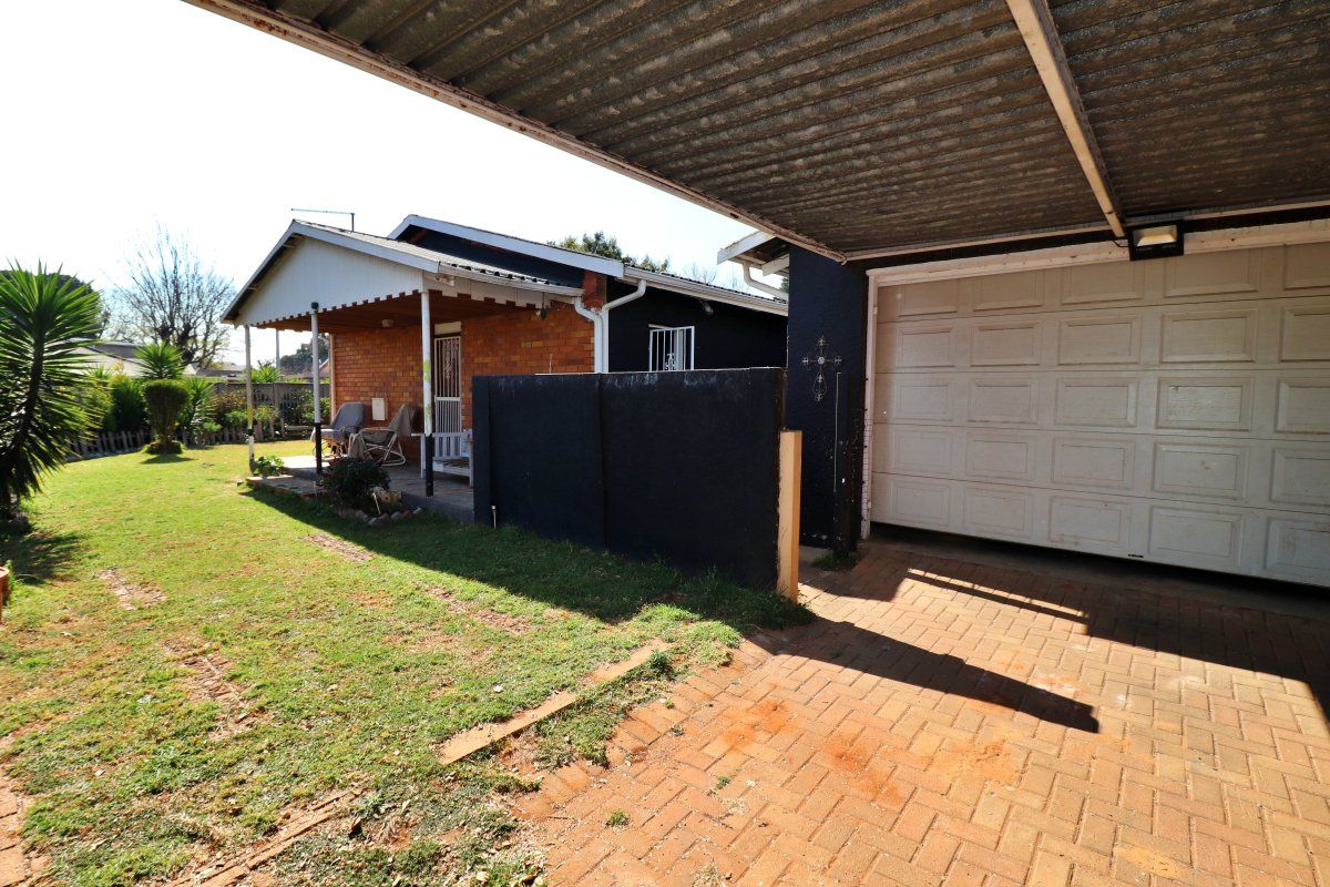Rear view of the property featuring a paved driveway, a covered carport, a garage, and a small covered patio overlooking the garden.