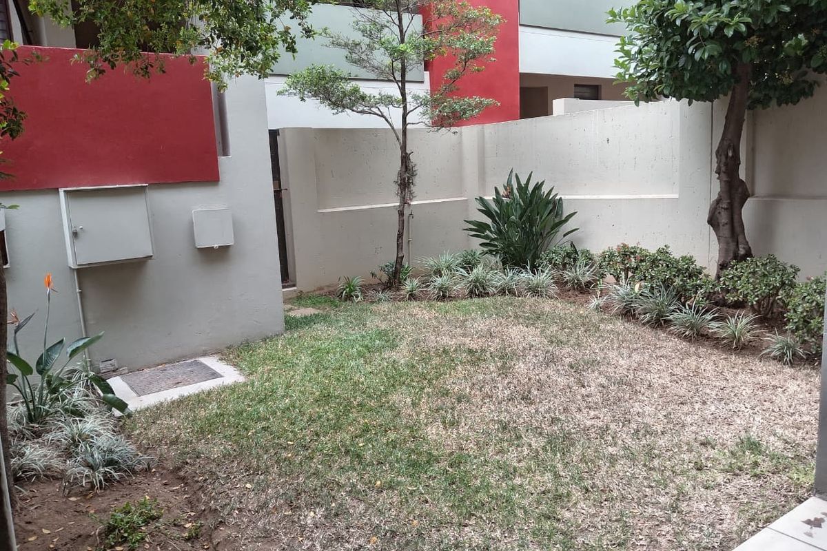 A private rear garden area featuring a boundary wall, established trees, and a lawn space within an apartment complex setting.