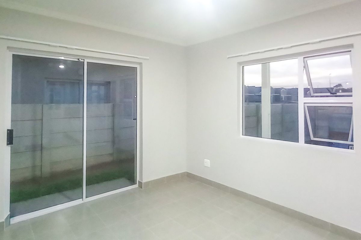 A bright, empty living room featuring neutral tiled flooring, a sliding glass door leading outside, and a large window providing natural light.