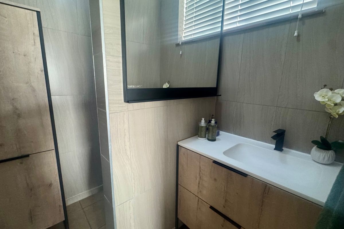 Modern bathroom featuring a wood-finish vanity with integrated basin, black fixtures, wall-mounted mirror, and neutral floor-to-ceiling wall tiles.