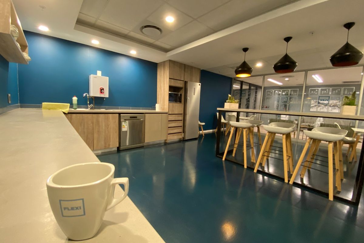 Modern commercial kitchen area featuring built-in cabinetry, integrated appliances, sleek epoxy flooring, and a breakfast bar with pendant lighting, adjacent to a glass-partitioned office space.