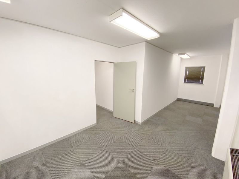 A clean, vacant office space featuring neutral walls, patterned carpet tiles, and functional overhead fluorescent lighting.