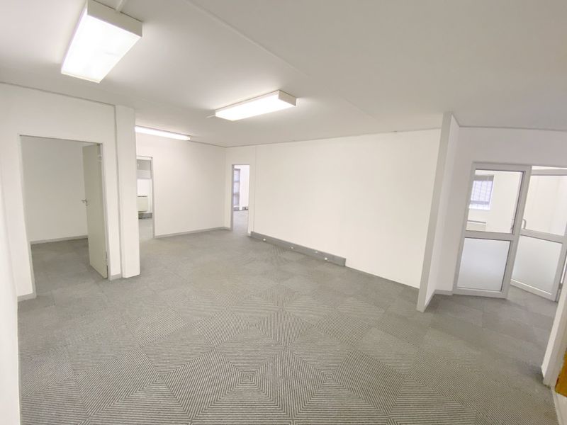 A clean, vacant commercial office space featuring neutral carpet tiles, bright fluorescent lighting, and multiple internal access doors.