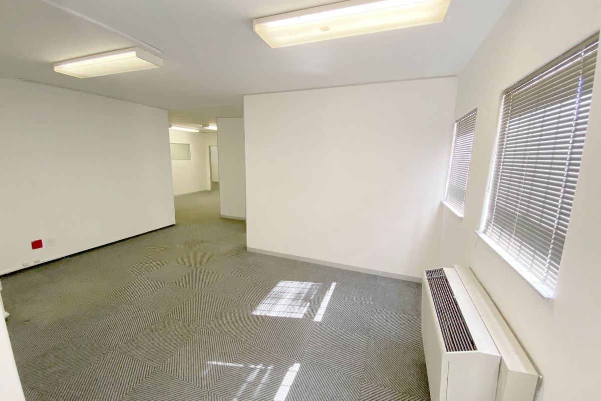 A clean, vacant commercial office space featuring carpeted flooring, built-in HVAC unit, and large windows with fitted venetian blinds.